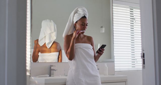 Woman multitasking by brushing her teeth while using smartphone in bathroom. Suitable for themes around morning routines, personal care, hygiene, and modern lifestyle. Ideal for websites, blogs, or advertisements promoting bathroom products, dental care, or smartphone usage.