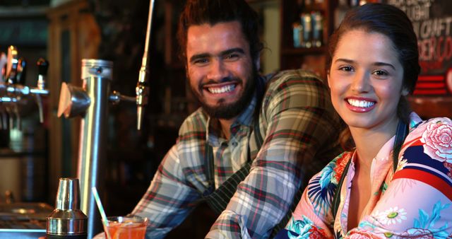 Portrait of smiling barmaid and barman with cocktail shaker and cocktail at bar counter in pub