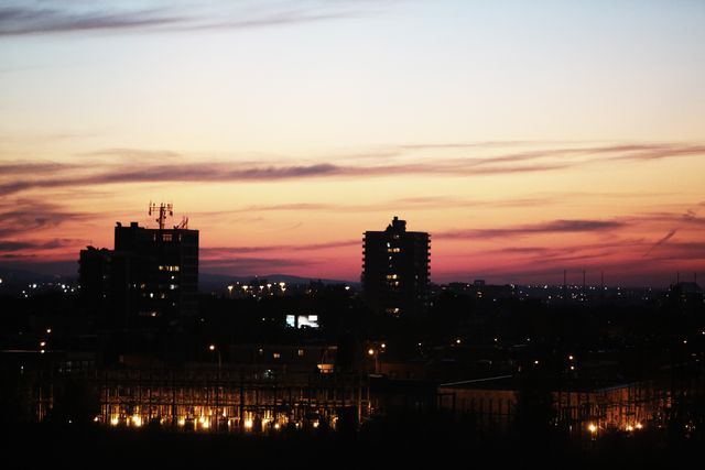 Urban skyline features dark silhouettes of buildings against an orange twilight sky. Ideal for themes related to modern life, evening, city landscapes, and urban settling.