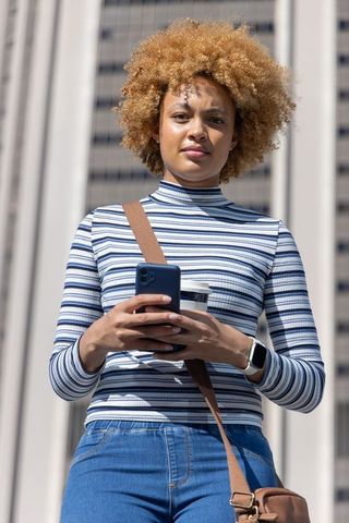 Confident african american woman standing by office tower holding smartphone and coffee