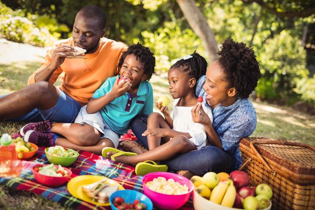Family enjoying a picnic in a park on a sunny day. Parents and children sitting on blanket, eating fruits, and talking. Ideal for use in advertisements, family-oriented promotions, health and wellness campaigns, and outdoor activity brochures.