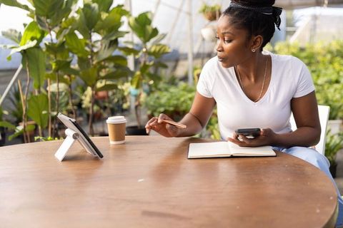 African american woman working remotely in greenhouse using tablet, phone and notebook