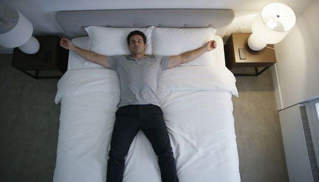 Man lying starfish on king bed relaxing in minimalist bedroom with warm bedside lamps