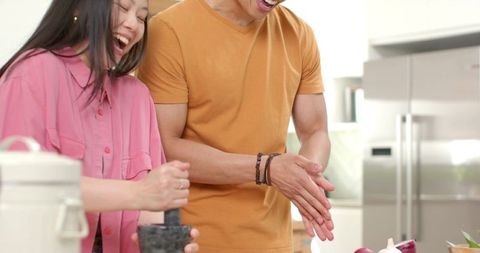 Multicultural couple laughing and preparing food in modern kitchen using mortar and pestle