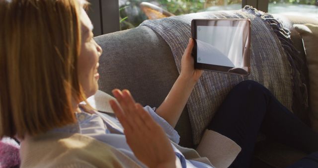 Woman relaxing on sofa using tablet for video call. Great for concepts of remote communication, technology in daily life, home comfort, digital interaction, and casual modern living.