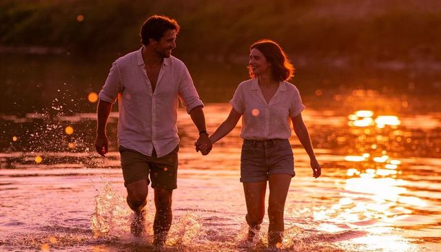 Couple wading hand in hand at golden hour sunset, laughing and splashing on backlit shoreline