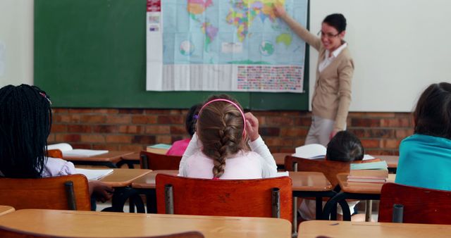 The teacher is standing at the front of a classroom, pointing at a world map on the board, while several seated children are watching attentively. This could be used for educational content, teaching materials, school-related articles, back-to-school campaigns, or blogs discussing effective teaching techniques and classroom engagement.
