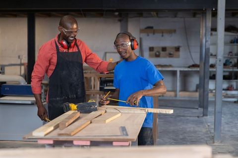 African american mentor teaching apprentice measuring wood at carpentry workbench