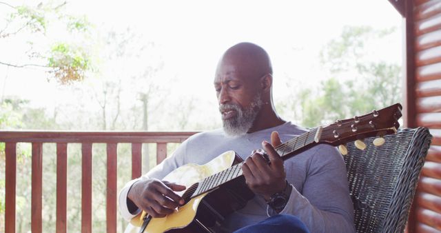 Elderly man with grey beard playing acoustic guitar on porch, enjoying a peaceful moment of music. Ideal for emphasizing relaxation, leisure, senior living, passion for music, outdoor hobbies, retirement activities, and serene settings. Suitable for promoting lifestyle blogs, wellness products, and health-related campaigns.