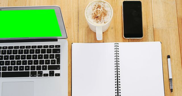 A laptop with a green screen sits next to a cup of coffee, a smartphone, and an open notebook with a pen on a wooden desk, with copy space. Ideal for a technology or work-from-home concept, the setup suggests a productive environment for business or creative tasks.