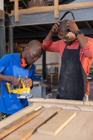 African american male carpenters cutting wood and collaborating with jigsaw in busy workshop