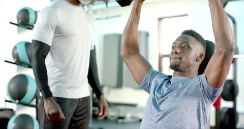 African american trainer spotting athlete performing seated overhead dumbbell press in sunlit gym