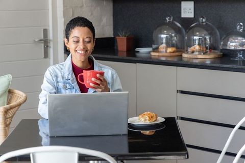 African american woman working from home in kitchen holding red mug, smiling at laptop