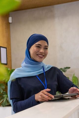 Asian woman receptionist wearing blue hijab smiling at clinic reception holding tablet