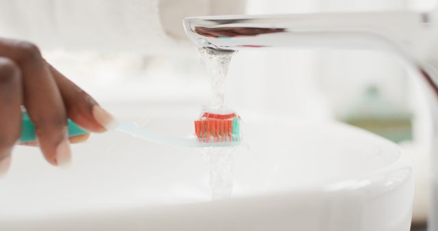 Close-up of a person's hand holding a toothbrush under running water from a faucet in a bright bathroom. Suitable for illustrating themes related to daily hygiene, dental care, morning routines, cleanliness, and personal health habits.
