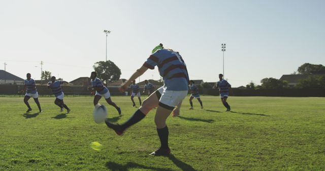 Rugby player in action kicking ball on a sunny field with teammates and opponents in the background. Ideal for use in sports articles, advertisements for athletic gear, team spirit promotions, and content highlighting physical fitness and teamwork.
