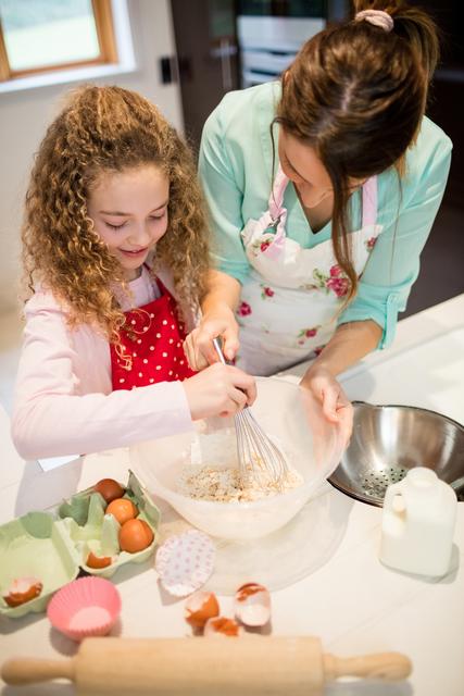 Mother assisting daughter with whisking flour in a kitchen, showcasing a moment of family bonding. Perfect for use in content about family activities, parenting, teaching children, cooking recipes, and promoting kitchenware products. Ideal for blogs, advertisements, and educational materials.