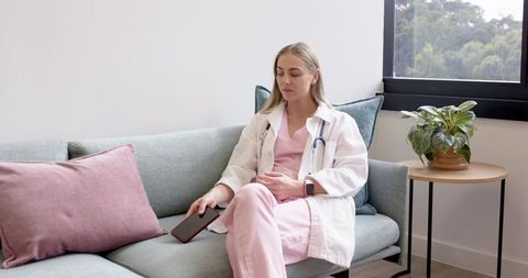 Female clinician in pink scrubs with stethoscope sitting on lounge sofa checking smartphone