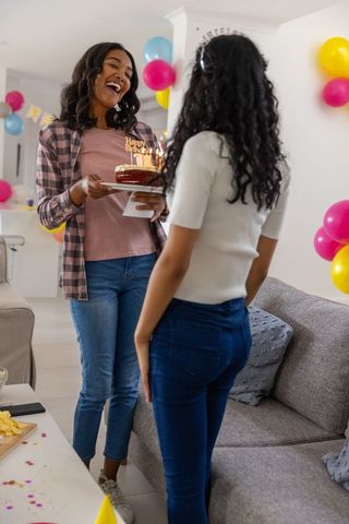 Sisters celebrating birthday at home holding lit cake with balloons and banner, smiling