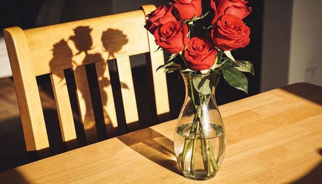 Sunlit red roses casting shadows on wooden table with glass vase and warm morning glow