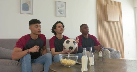 Watching soccer match together multicultural friends on couch holding ball and snacks