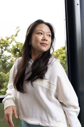 Asian woman on balcony leaning on railing wearing cream hoodie, contemplative portrait