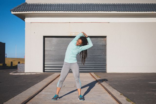 Biracial woman with dreadlocks stretching in front of a garage door, wearing sportswear. Ideal for promoting fitness, healthy lifestyle, outdoor exercise routines, and urban workout settings. Suitable for use in fitness blogs, health magazines, and sportswear advertisements.