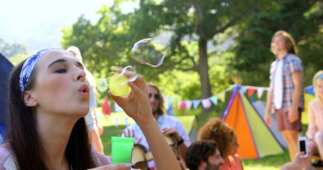Image depicts young woman blowing bubbles at an outdoor festival surrounded by friends. Colorful tents and festive decoration enhance joyful summer atmosphere. Perfect for promotional use in outdoor events advertisements, lifestyle blogs, social media content related to festivals and celebrations.