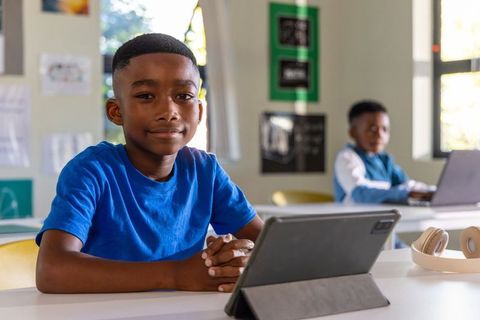 African American Preteen Using Tablet in Bright Classroom While Classmate Works on Laptop