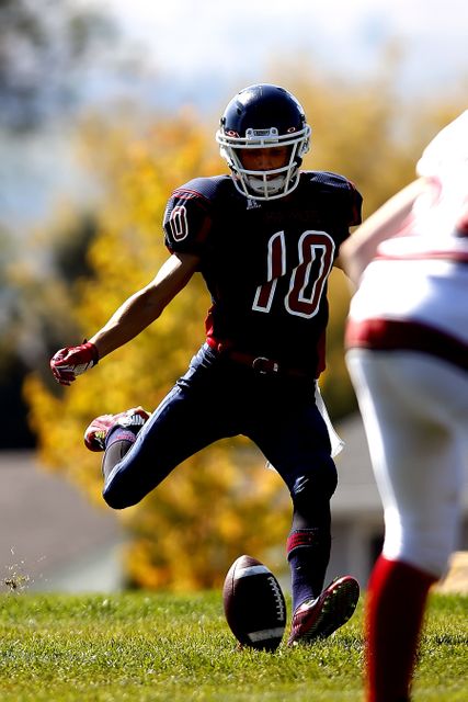 Youth football player kicking ball during a game on a sunny day outdoors. Ideal for illustrating children's participation in sports, teamwork, competitive spirit, and youth athletic programs. Can be used on sports education websites, youth football promotions, and community sports activity advertisements.
