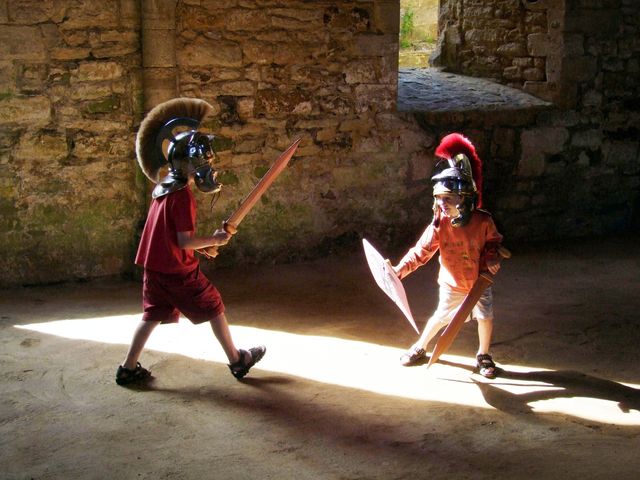 Young boys engaged in playful knight battle in ancient ruins, wearing helmets and wielding wooden swords and shields. Use for themes of imagination, historical play, medieval reenactments, and childhood fun.
