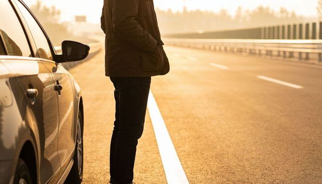 Man standing by silver sedan on empty highway shoulder at golden hour, backlit solitude