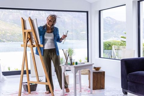 Mature woman painting at easel in bright lakeside studio wearing headphones, serene view