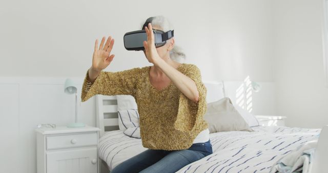 Senior woman using VR headset while sitting on bed at home. She is actively engaging with virtual environment using her hands. Perfect for illustrating aging and technology, modern lifestyle of elderly generation, innovation, and new experiences for seniors.