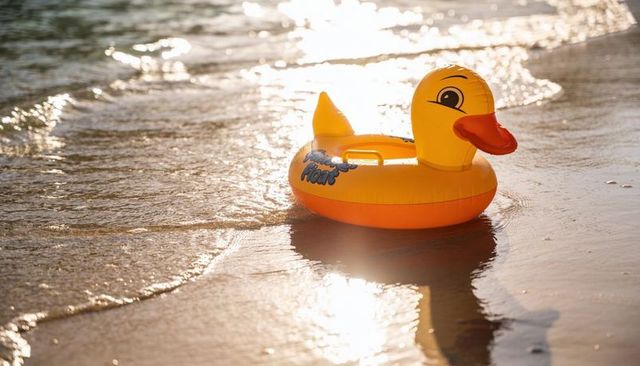 Sunlit yellow inflatable duck swim ring reflecting on wet sand at shoreline for summer