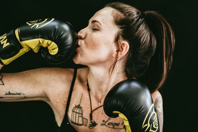 Female boxer enthusiastically kissing glove in celebration of a victorious moment. She has multiple tattoos and is wearing black and yellow boxing gloves. This energetic and empowering image can be used for promoting fitness programs, athletic apparel, women's empowerment campaigns, or sports-related content.