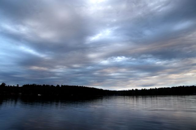 Serene lake reflecting a dramatic cloudy sky at dusk. Trees form a dark silhouette along the horizon. Ideal for backgrounds, nature themes, tranquility, relaxation, meditation, or environmental awareness campaigns.