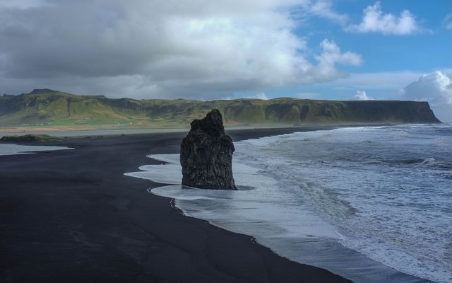 Stunning black sand beach featuring a prominent sea stack off the coast of Iceland. Dark volcanic sand contrasts beautifully with the white foamy waves of the ocean and the dramatic cloudy sky. Ideal for travel and nature articles, coastal landscapes, and promoting Icelandic tourism.