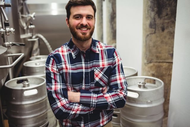 Bearded man in plaid shirt standing with arms crossed in brewery, smiling confidently. Stainless steel storage tanks visible in background. Ideal for use in articles about brewing industry, craft beer production, or workplace environments in manufacturing settings.