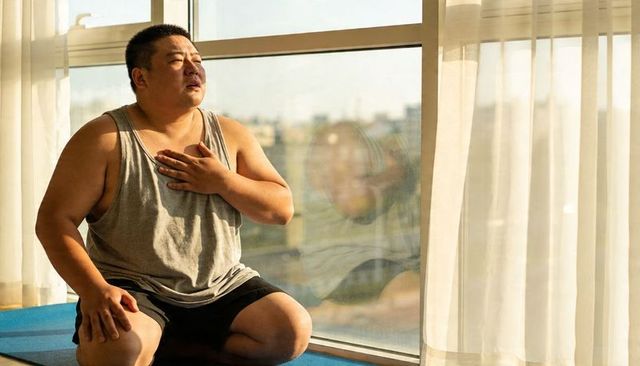 Man practicing deep breathing on blue mat by window in sunlit room with sheer curtains
