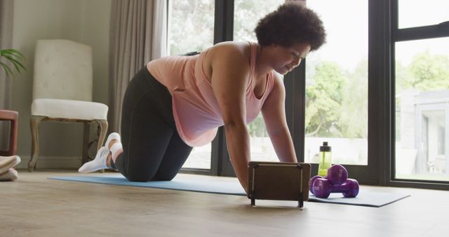 Woman is doing an exercise on a yoga mat in a living room setting with large window offering natural light. She has dumbbells and a water bottle beside her. Perfect for use in articles or advertisements promoting home fitness routines, health and wellness, and indoor exercises.