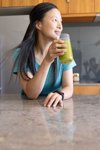 Asian woman enjoying green smoothie while checking smartwatch in modern kitchen
