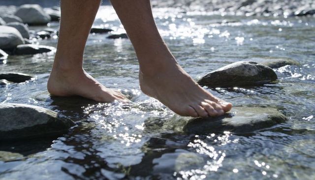 Barefoot stepping across sunlit rocky creek on smooth river stones lifestyle outdoors