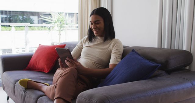Young woman sitting comfortably on a couch in a modern living room, using a tablet. This image can be used for themes related to technology, relaxation, home living, remote work, or casual lifestyle settings.