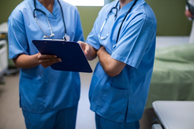 Healthcare professionals in blue scrubs are discussing patient records in a hospital ward. This image can be used to depict teamwork, patient care, and medical discussions in healthcare settings. Ideal for use in medical articles, healthcare websites, and educational materials related to nursing and hospital environments.