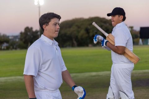 Cricketers practicing at dusk wearing white uniforms holding bats and gloves on outfield