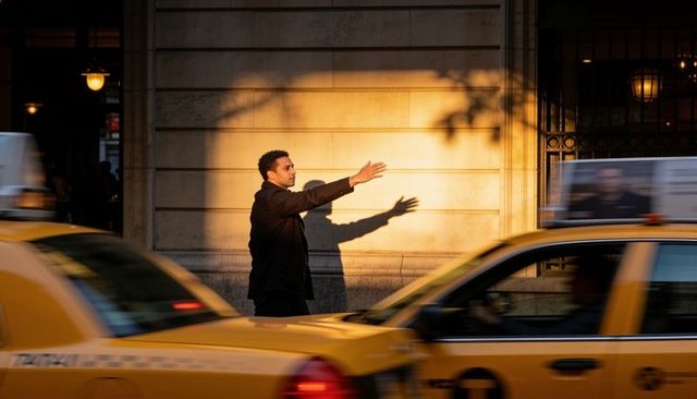 Man hailing taxi on sunlit urban sidewalk casting dramatic long shadow golden hour traffic motion
