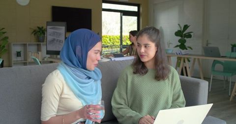 Diverse coworkers chatting on sofa, hijab woman holding glass, laptop teamwork in coworking space