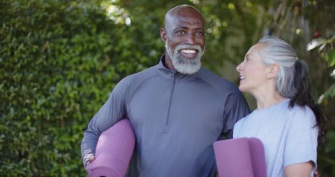 Active senior interracial couple carrying yoga mats, walking and smiling in sunlit park
