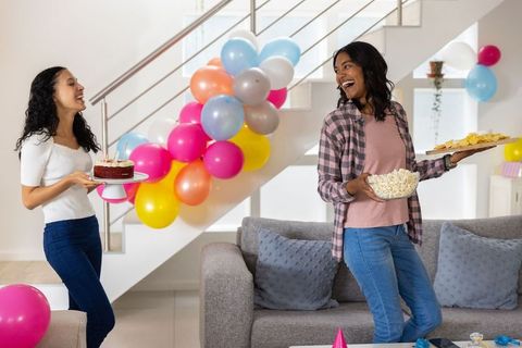Diverse sisters celebrating birthday home carrying chocolate cake balloons snacks laughing